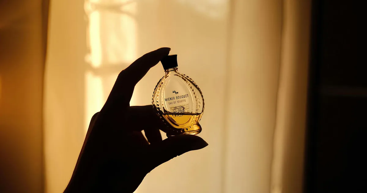 A silhouette of an elegant perfume bottle held by a hand against a warm, dimly lit background.