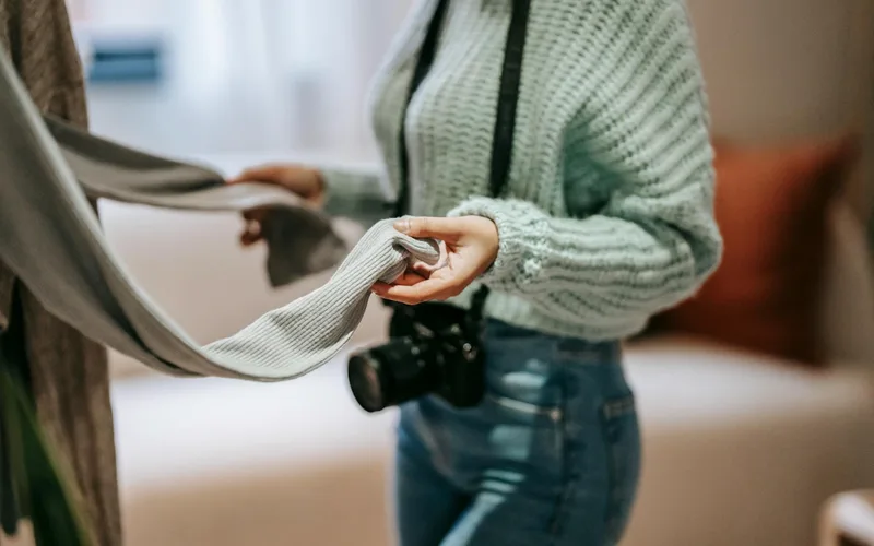 Woman in green sweater holding fabric, a camera hanging, in a cozy living room setting.