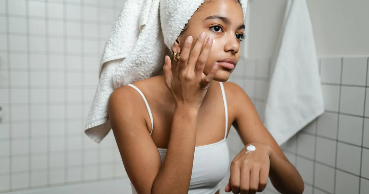 A young woman in a bathroom applying skincare cream with a towel on her head, focusing on self-care.