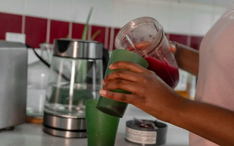 Close-up of a person pouring a healthy red smoothie into a green glass in a home kitchen.