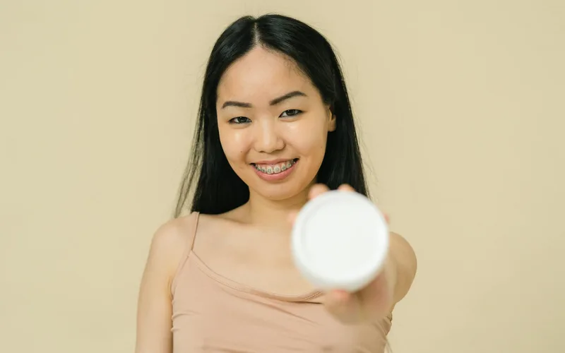 Portrait of a young woman with braces holding a cosmetic product jar with a smile.