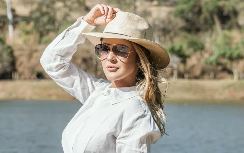 Elegantly dressed woman in hat and sunglasses posing by the water on a sunny day.