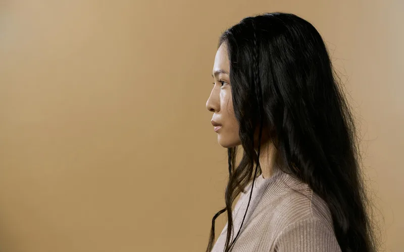 A side profile portrait of a woman in a sweater against a neutral beige background.