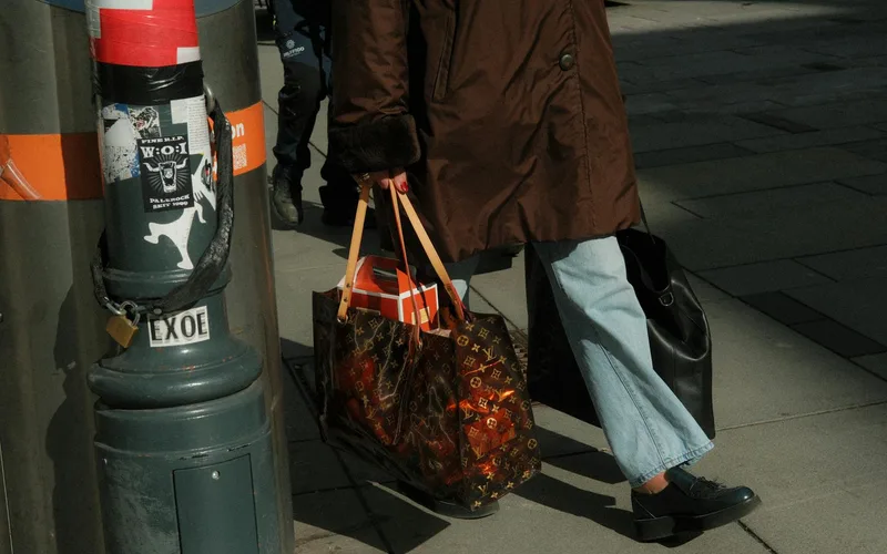 A person in stylish attire walks with luxury brand shopping bags on a city street.