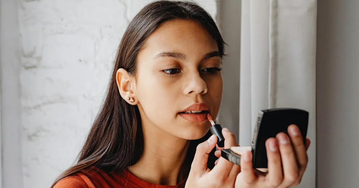 Young woman applying lip tint using a compact mirror indoors by a window.