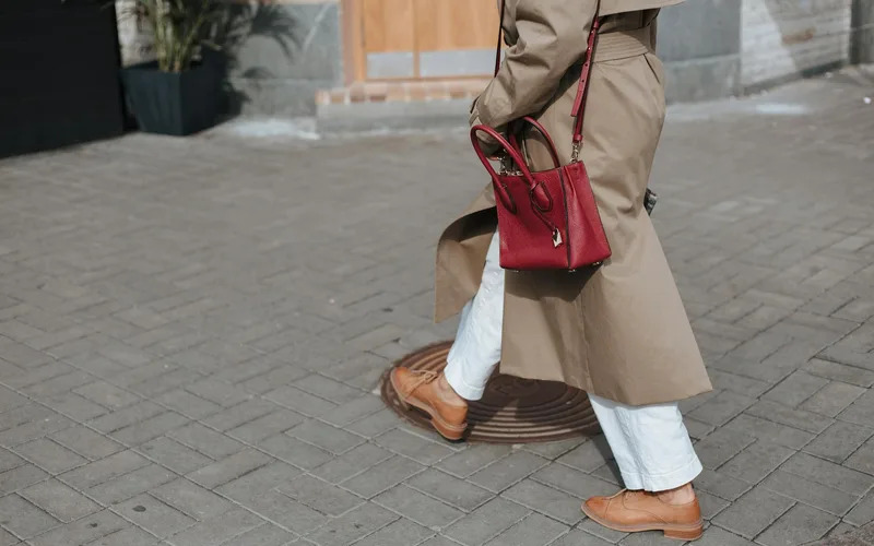 Person walking on pavement wearing a beige trench coat and holding a red handbag.