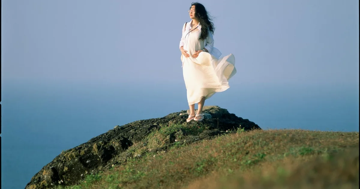Woman in a flowing white dress standing gracefully on a hilltop, overlooking the sea.