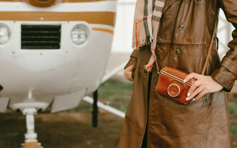 Stylish leather trench coat with cross body bag beside a small airplane outdoors.