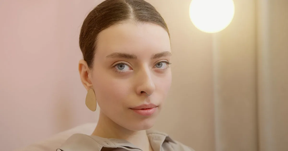 Portrait of a young woman with blue eyes, wearing earrings and a nose piercing, indoors with soft lighting.