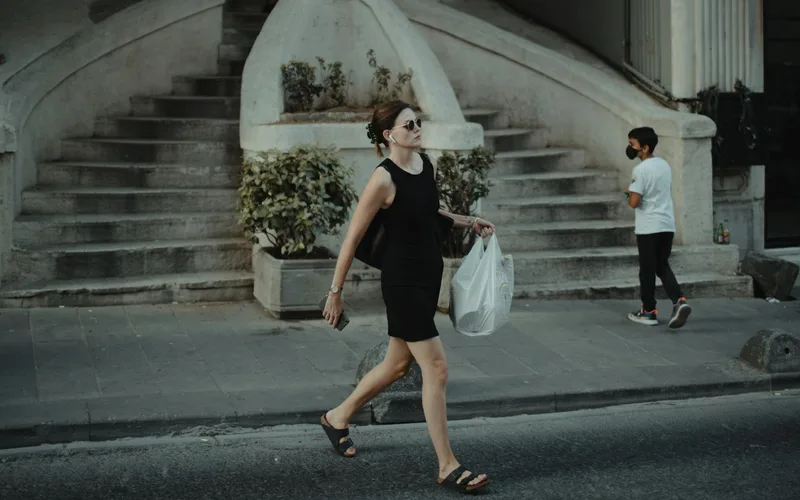 A woman in sunglasses and a black dress walks on the street carrying a white shopping bag.