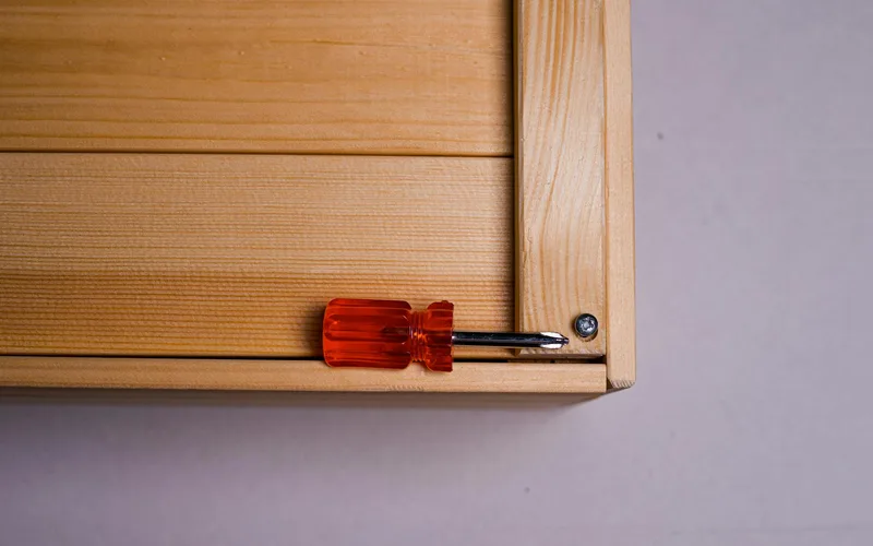 Red-handled screwdriver resting on a wooden surface corner with a visible screw.