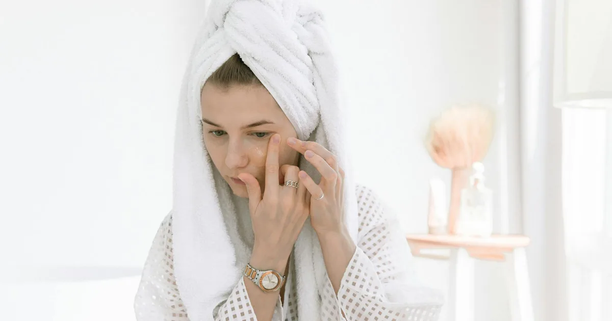 A woman in a bathrobe applies skincare cream in a bright, relaxing indoor setting.