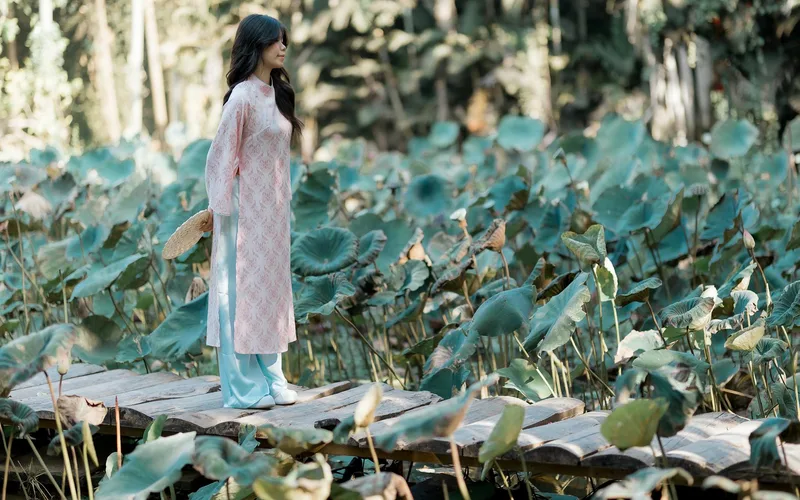 A woman in traditional attire standing on a rustic wooden bridge in a lush lotus garden.