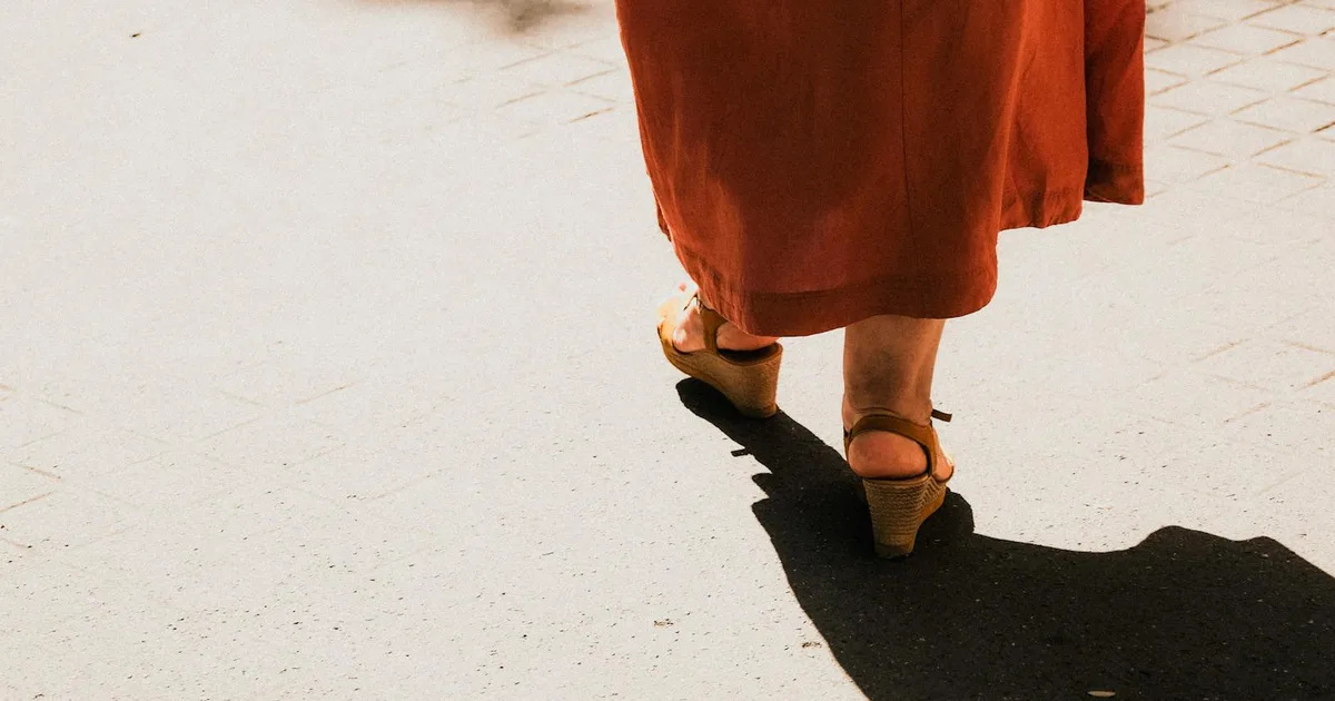 Close-up of a woman in a dress and heels walking on a sunlit urban sidewalk.