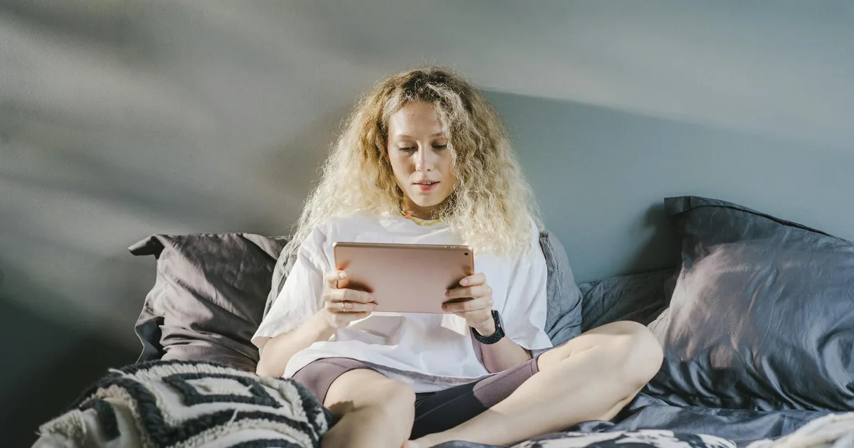 A woman with blonde hair sits on a cozy bed using a digital tablet indoors.