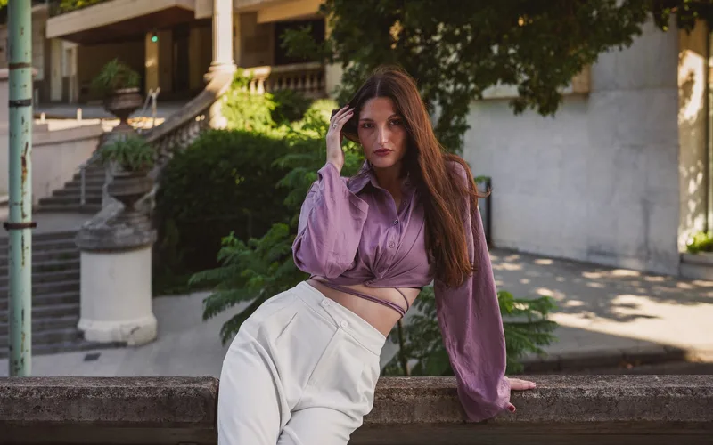 Fashionable woman in a purple blouse poses elegantly outdoors with architectural backdrop.