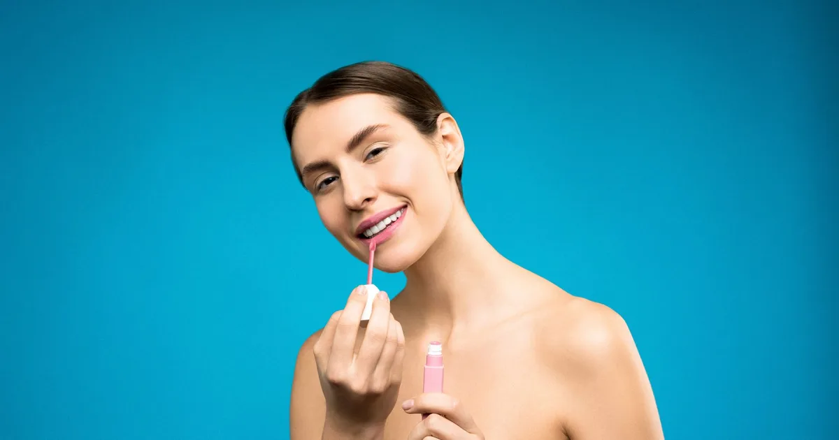 A smiling woman applies pink lipstick against a vibrant blue background, showcasing beauty and confidence.