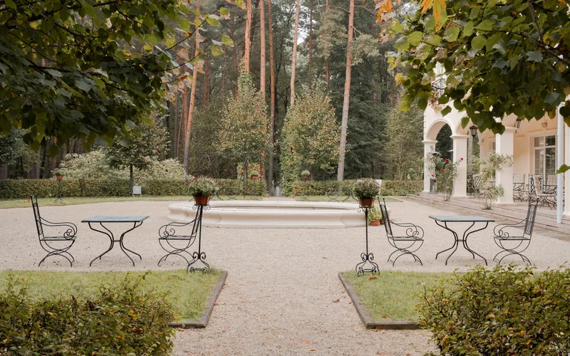Serene garden courtyard with iron furniture surrounded by lush green trees and mansion architecture.