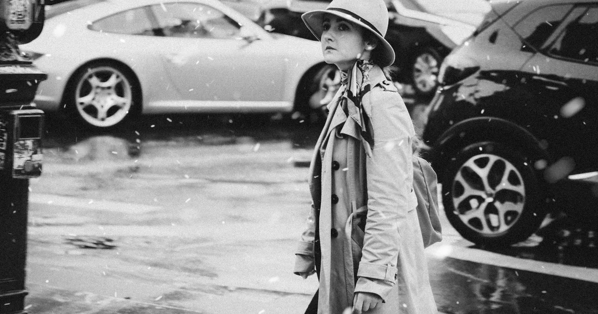 Black and white photo of a woman in outerwear walking in a snowy street with vintage cars.