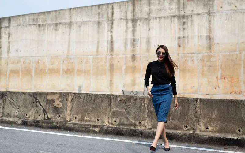Stylish woman in denim skirt strolling down a city street with concrete wall background.