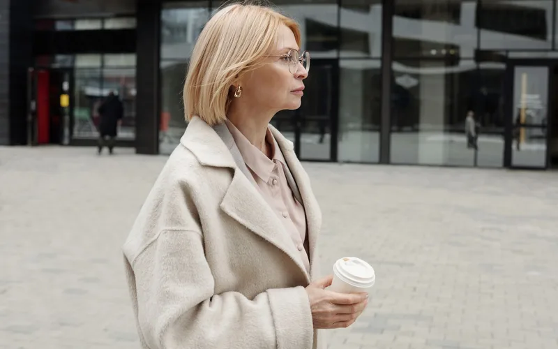 Woman in a beige coat holding coffee, standing confidently in an urban setting.