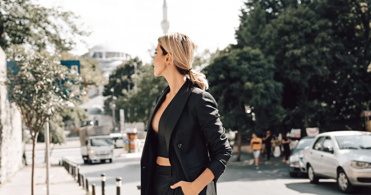 Elegant female in trendy outfit with hand in pocket standing on pavement near road with driving cars