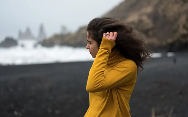 Profile of a woman with long hair in a yellow sweater on a windy rocky shore.