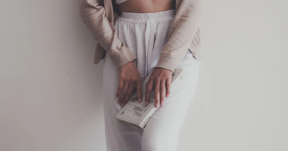 Stylish woman in beige cardigan and white crop top holding a book against a white background.