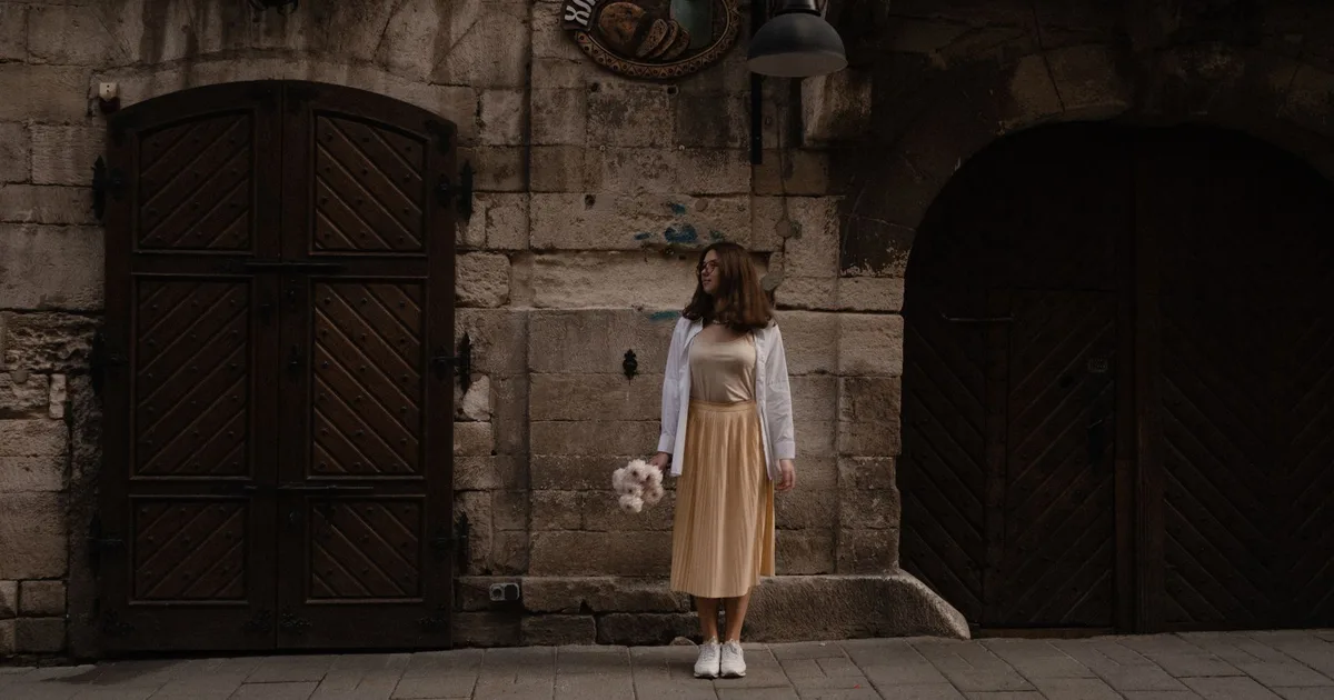 Elegant woman with flowers stands by rustic wooden doors on a historic street.