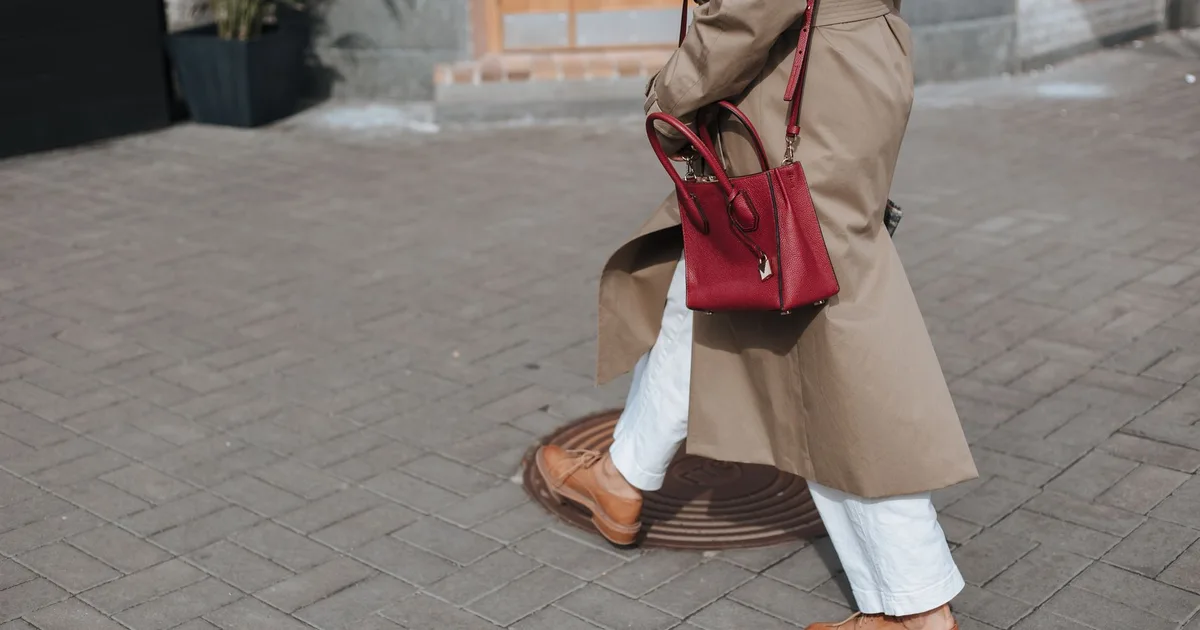 Person walking on pavement wearing a beige trench coat and holding a red handbag.
