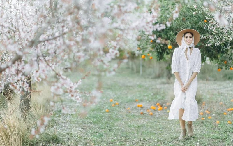 A woman in a white dress and hat stands amidst blooming cherry blossoms and fallen oranges, exuding spring elegance.