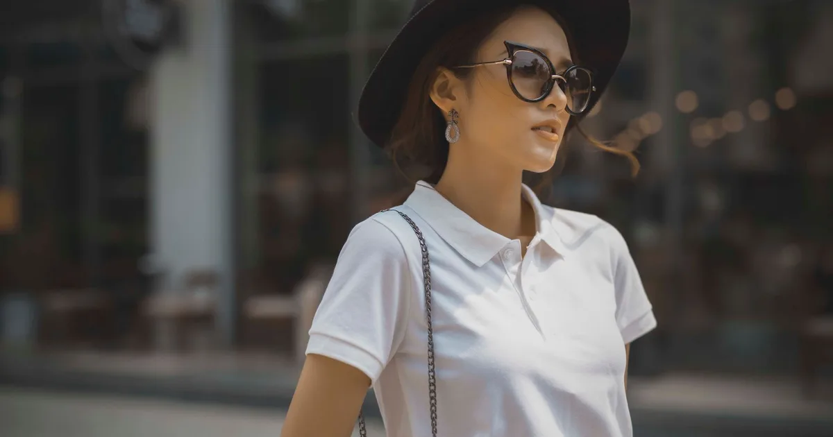 Fashionable woman in a white polo shirt, sunglasses, and black hat outdoors, exuding style and confidence.