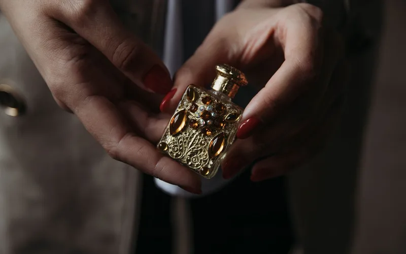 Crop faceless female with red manicure demonstrating small vintage golden perfume bottle decorated with natural stones and carved details