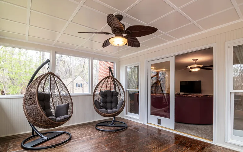 Bright sunroom featuring stylish egg chairs, ceiling fan, and wooden flooring with large glass windows.