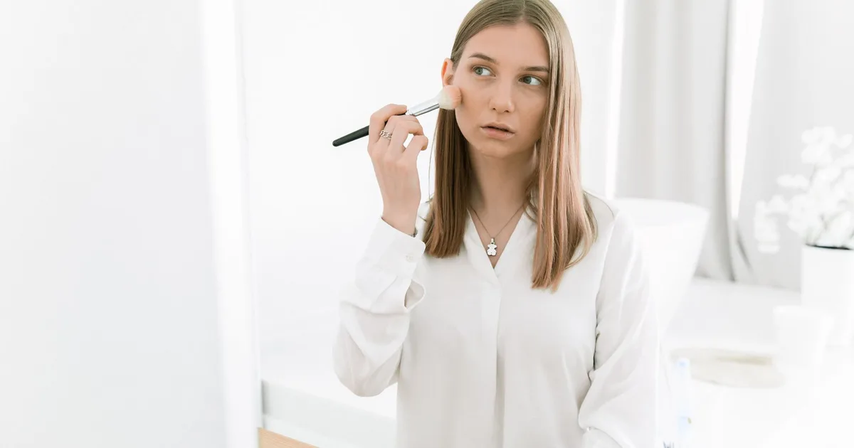A woman wearing white long sleeves applies blush indoors using a makeup brush.