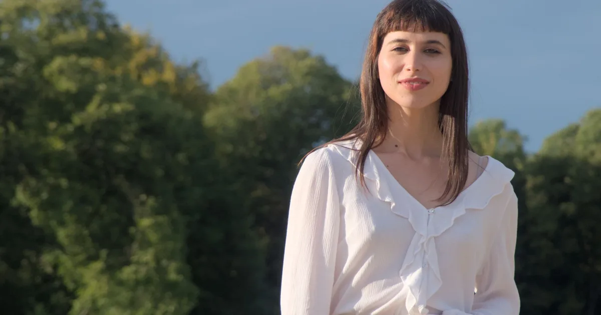 A young woman wearing a ruffled white blouse poses confidently in a park setting.