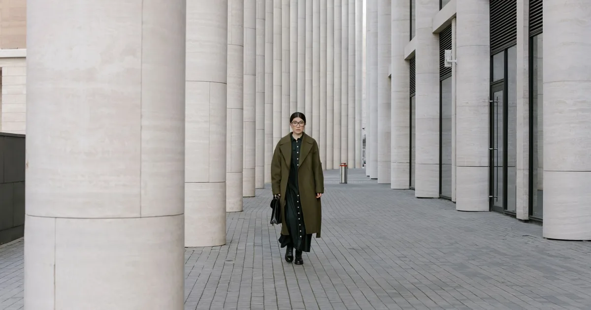 Asian woman in a long coat walking confidently through a modern architectural space with tall columns.