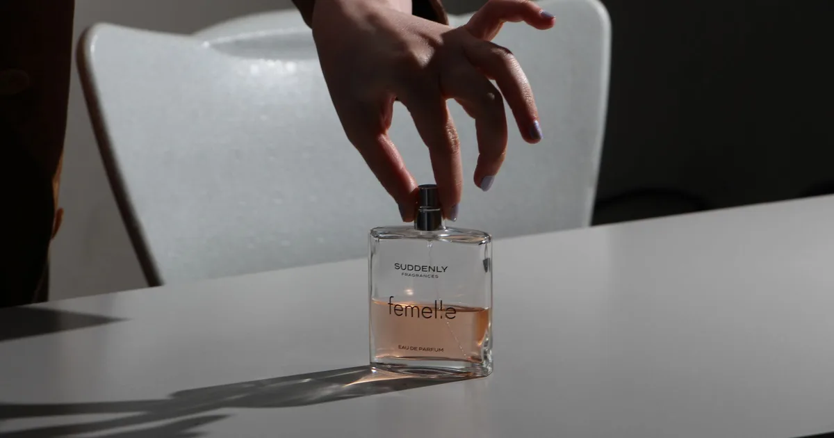 A hand with manicured nails gracefully holds a perfume bottle on a table in soft lighting.