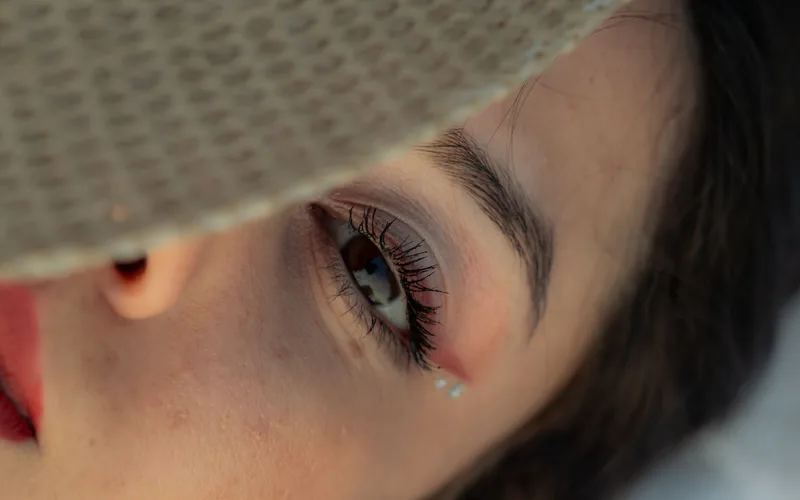 Detailed close-up of a woman's eye shaded by a sun hat, featuring makeup and jewels.
