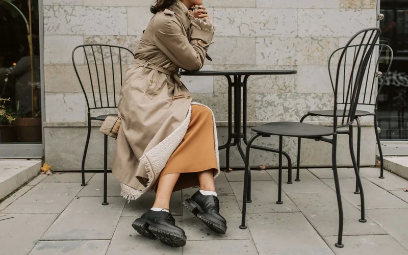 Stylish woman in a trench coat and black shoes at an outdoor cafe table.