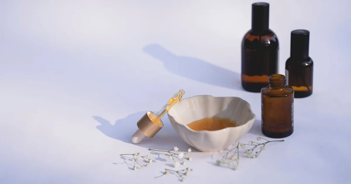 Elegant minimalistic display of amber glass bottles, ceramic bowl, dropper, and flowers on white background.