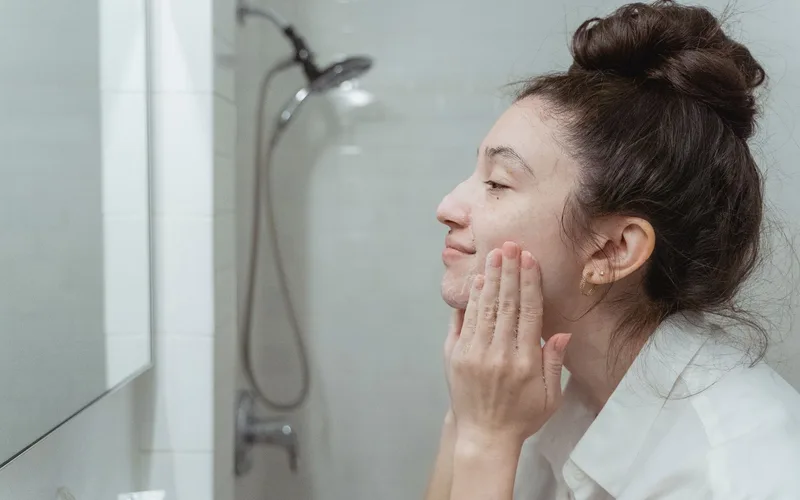 A woman applying facial cream in a mirror for her skincare routine in a bathroom.