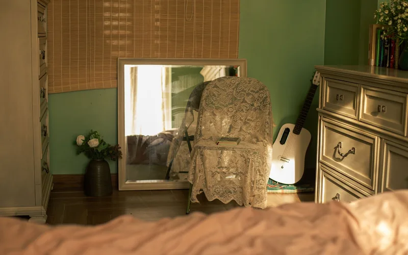 Inviting bedroom corner featuring a white guitar, lace draped chair, and floral arrangements, bathed in warm sunlight.