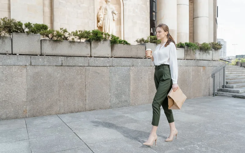 Confident woman in stylish attire walks outdoors holding coffee and documents.